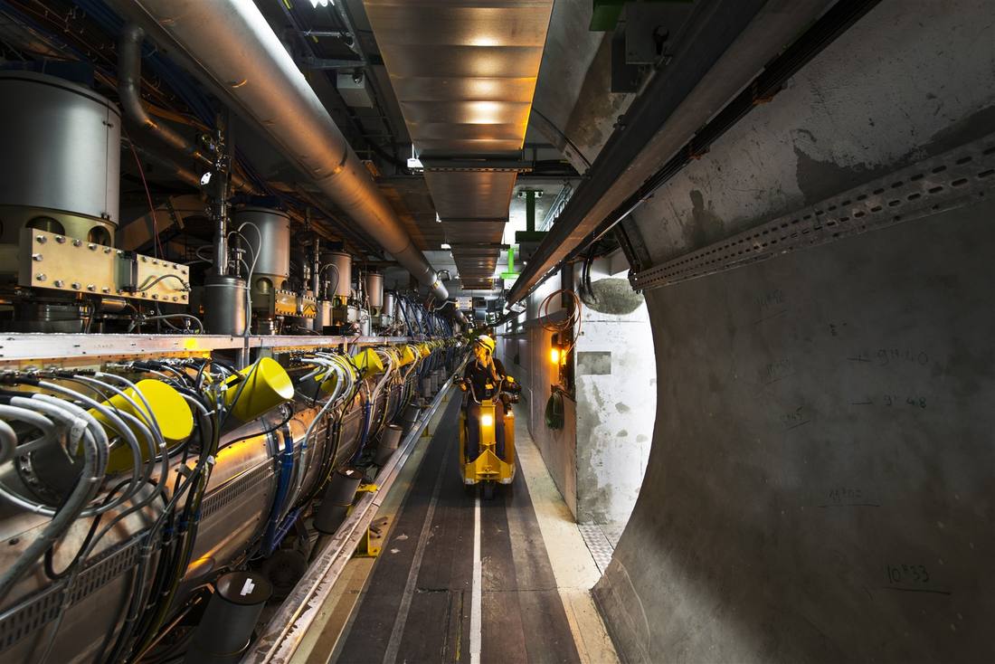 A technician riding a small cart through the LHC tunnel alongside a row of magnets and cabling A technician riding a small cart through the LHC tunnel alongside a row of magnets and cabling